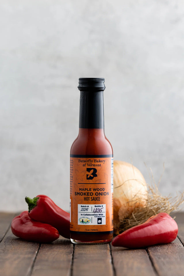 a bottle of smoked onion hot sauce on a dark wood table surrounded by red serrano and jalapeño peppers with a large onion behind it