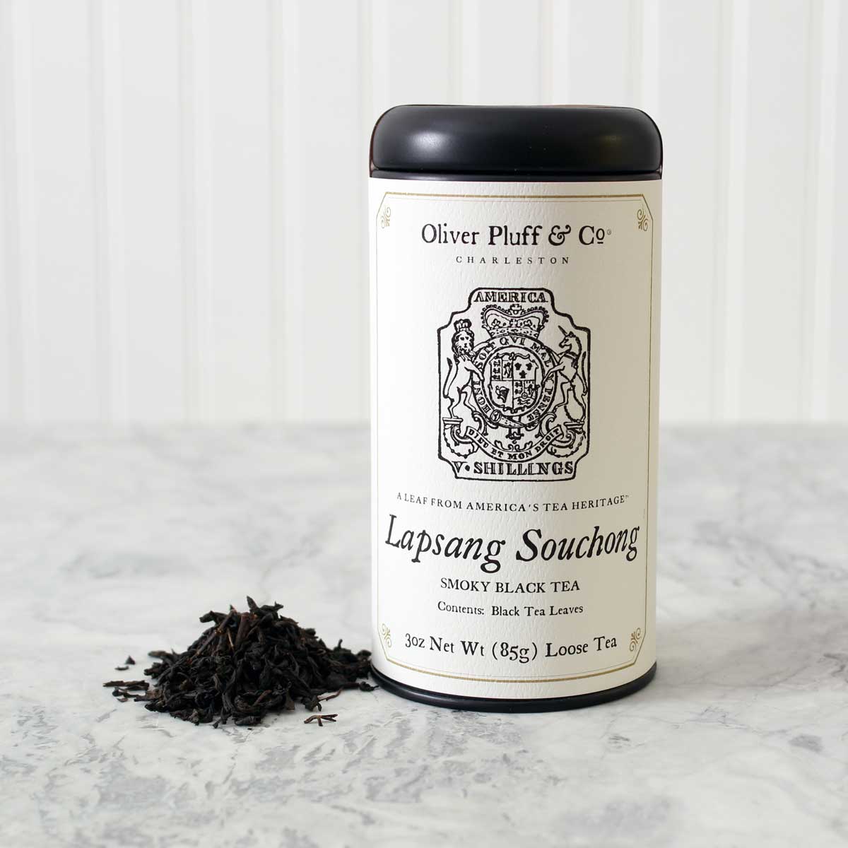 a tin of Lapsang Souchong loose tea with matte black tin and white printed label with image of a tax stamp from the 1765 stamp act on a white marble surface with white background