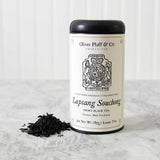 a tin of Lapsang Souchong loose tea with matte black tin and white printed label with image of a tax stamp from the 1765 stamp act on a white marble surface with white background