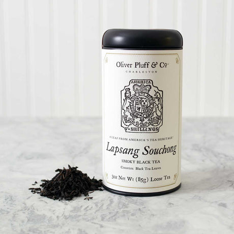 a tin of Lapsang Souchong loose tea with matte black tin and white printed label with image of a tax stamp from the 1765 stamp act on a white marble surface with white background