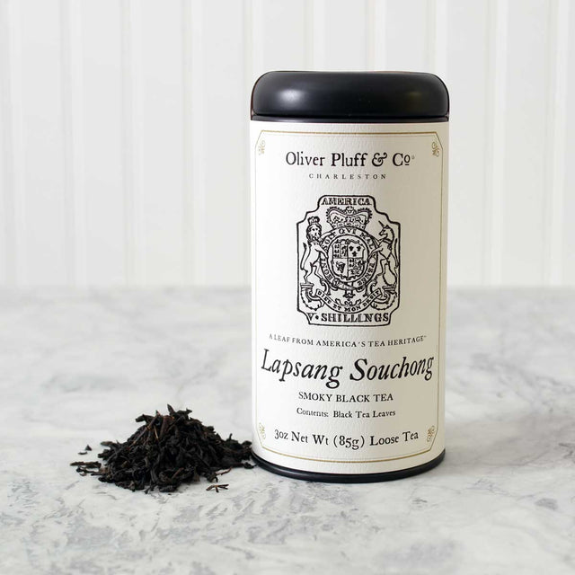 a tin of Lapsang Souchong loose tea with matte black tin and white printed label with image of a tax stamp from the 1765 stamp act on a white marble surface with white background
