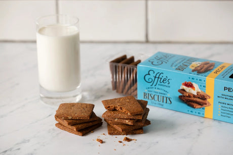 a marble countertop with a glass of milk next to an open box of effie's pecan biscuits stacked and ready to be dunked into milk