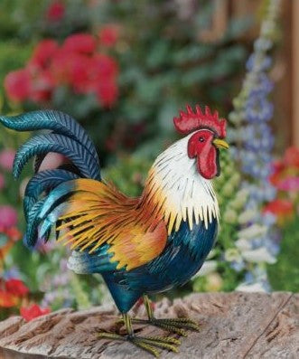 image of a life-size, metal, Golden Duckwing Rooster with golden wing feathers, white neck feathers, and red head details on a rustic, wooden table against a backdrop of a lush garden of flowers