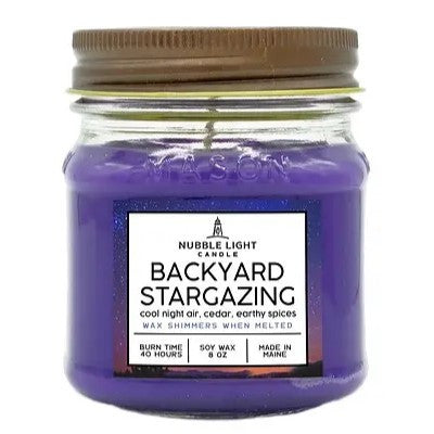 a candle in a clear glass mason jar with purple wax and a label that reads "Backyard Stargazing" against a white background