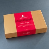 A rectangular brown cardboard box for Harbor Sweets' Chocolate Company, featuring a wide red paper band wrapped around the center with white text reading 'Sweet Loops' and product details, photographed on a dark gray surface.