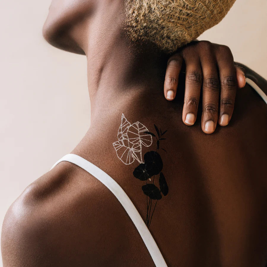 a model's upper back showing the tattoos in daylight - the boldness of the black plant tattoos and the white line of the glowing tattoos contrast beautifully on her deep skin tone.