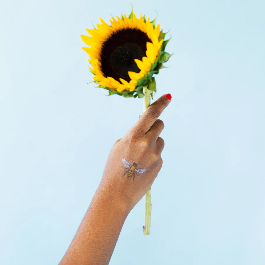 an outstretched hand holding a sunflower with a honey bee temporary tattoo on the model's wrist against a sky blue background